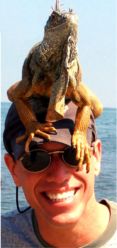 Smiling man wearing sunglasses with a large iguana perched on his hat beside the water.